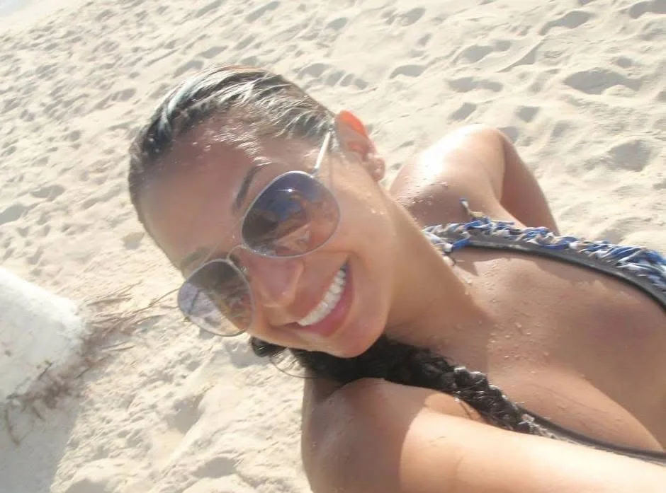 A smiling woman wearing aviator sunglasses and swimwear reclines on a sunny white sand beach.