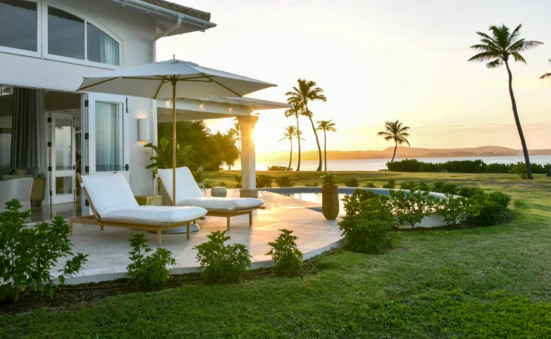 White sun loungers sit beside a private villa pool overlooking palm trees and a calm ocean at sunset.