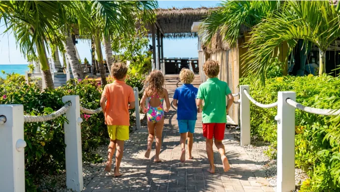 Four children in colorful swimwear run down a paved pathway lined with palm trees toward a thatched beach pavilion.