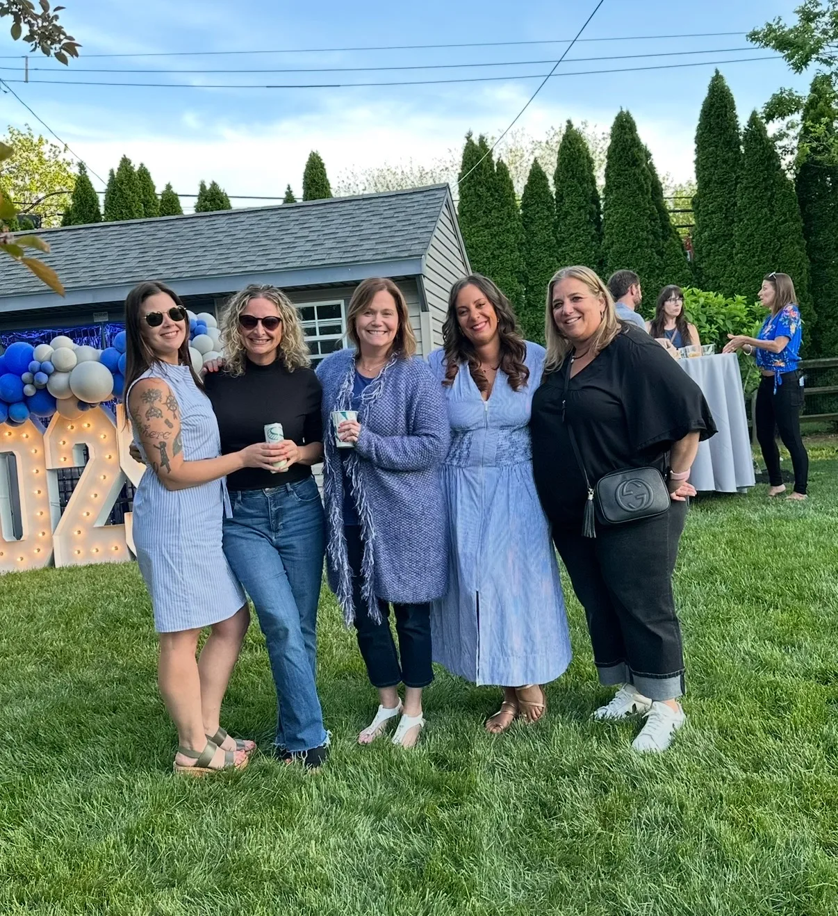 Five smiling women pose together on a green lawn during an outdoor celebration with tall evergreen trees in the background.