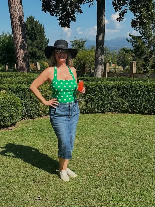 A woman in a wide-brim hat holds a cocktail, standing on a manicured garden lawn backed by hedges and trees.