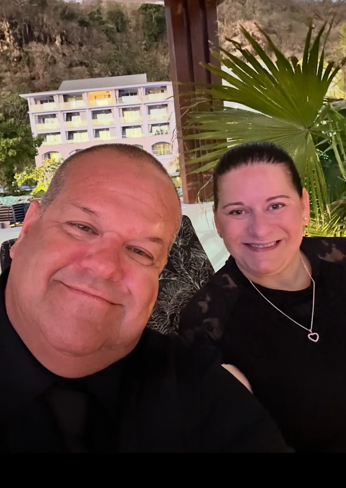 A smiling couple poses together on an outdoor terrace with tropical foliage and a multistory resort building in the background.