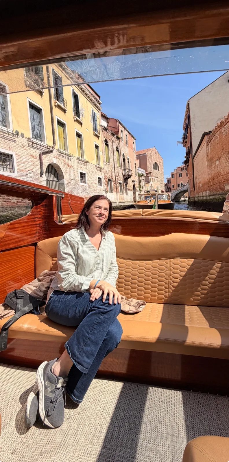 A woman sits aboard a wooden water taxi cruising along a sunlit canal lined with historic brick buildings in Venice.