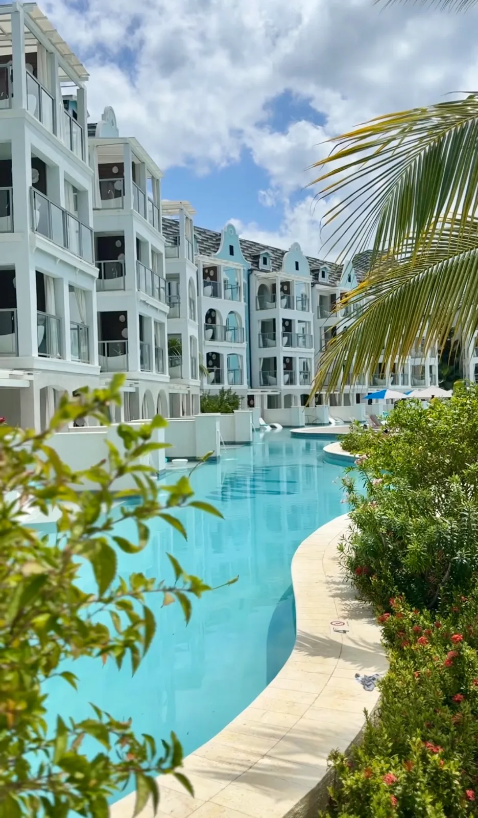 A turquoise swim-up lagoon pool curves past white multi-story resort suites bordered by tropical palms and lush greenery in Barbados.