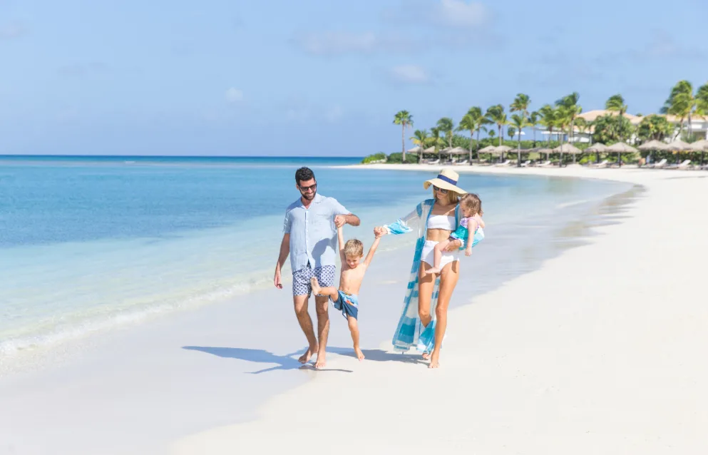 A family of four walks along a white sand beach beside calm turquoise ocean water lined with palm trees.