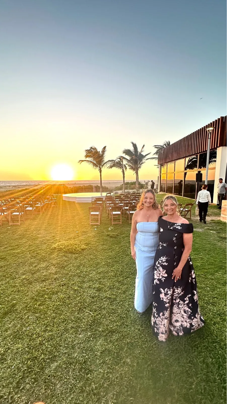 Two women in formal gowns stand on a coastal lawn prepared for an outdoor wedding ceremony at sunset.