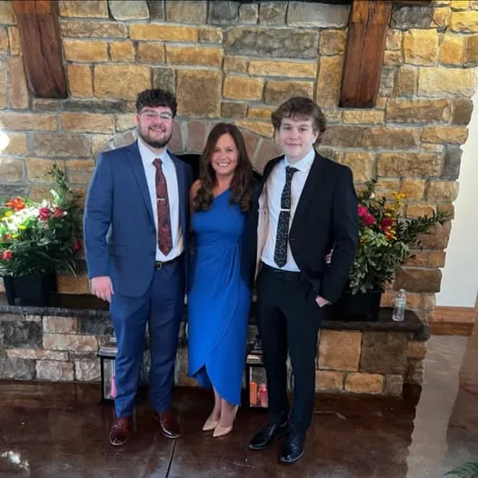 A woman in a vibrant blue dress stands between two men in formal suits before a rustic stone fireplace.