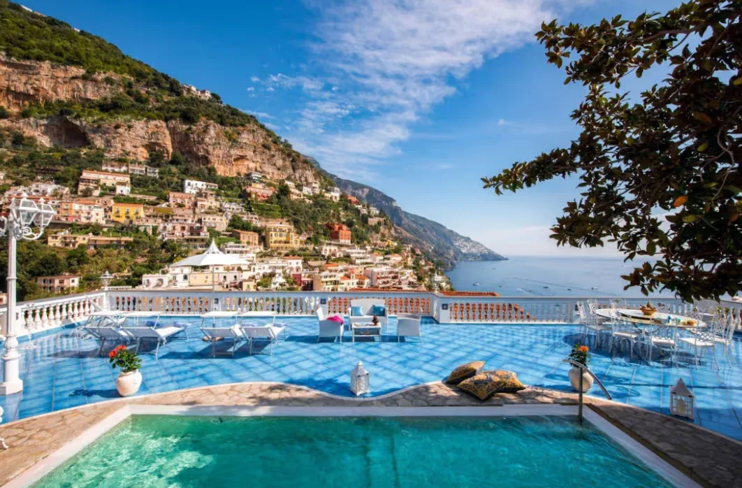 A swimming pool and blue-tiled terrace overlook colorful cliffside villas and the Mediterranean Sea in Positano, Italy.