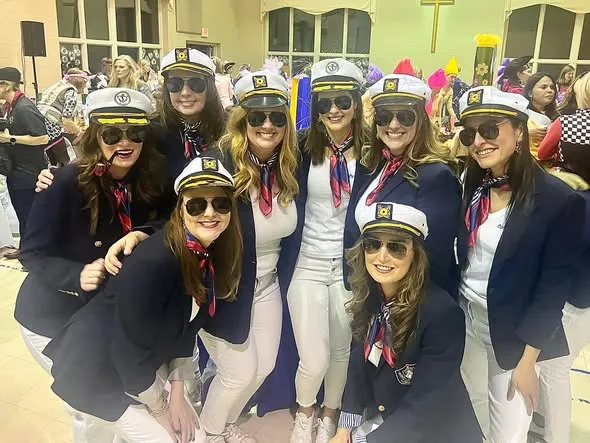 A group of women wearing matching captain hats, navy blazers, and sunglasses pose together at an indoor themed party.