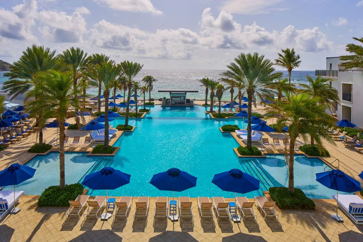A large resort swimming pool lined with palm trees, sun loungers, and blue umbrellas overlooks the ocean in Sint Maarten.