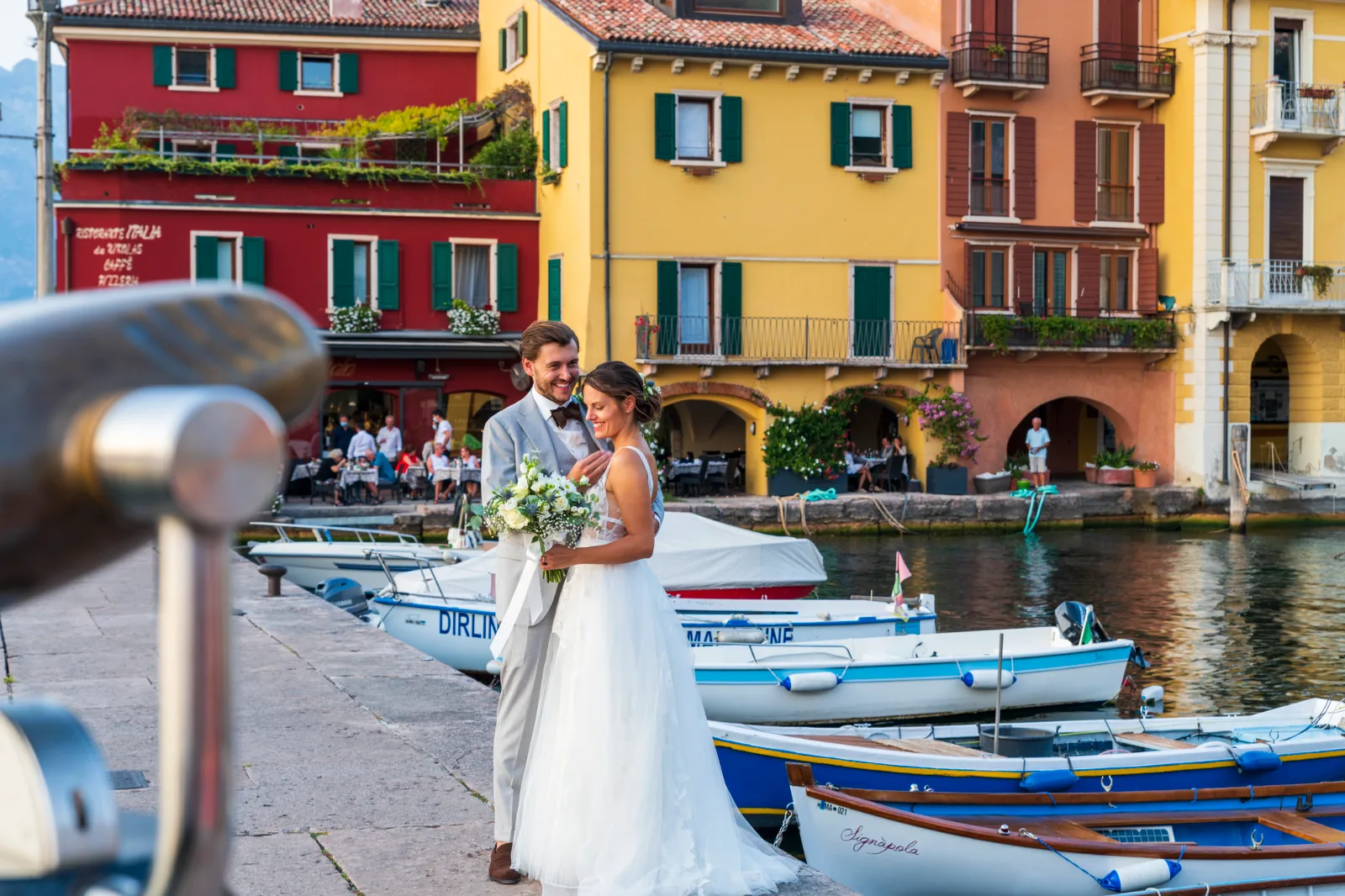 A smiling bride and groom stand together on a stone quay beside moored boats and colorful Italian buildings.