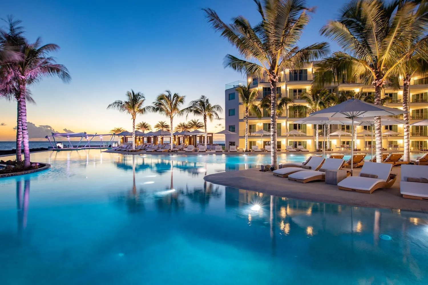 Illuminated swimming pool surrounded by palm trees and loungers beside a beachfront resort at dusk in St. Martin.