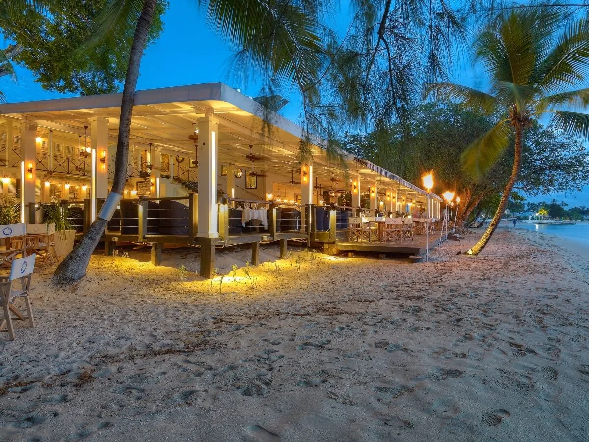 An open-air restaurant deck warmly illuminated at twilight sits directly on a sandy beach lined with palm trees in Barbados.