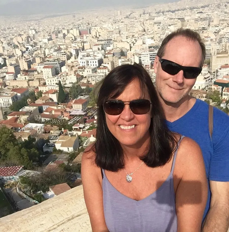 A smiling couple wearing sunglasses poses together on an overlook above the sprawling white cityscape of Athens, Greece.
