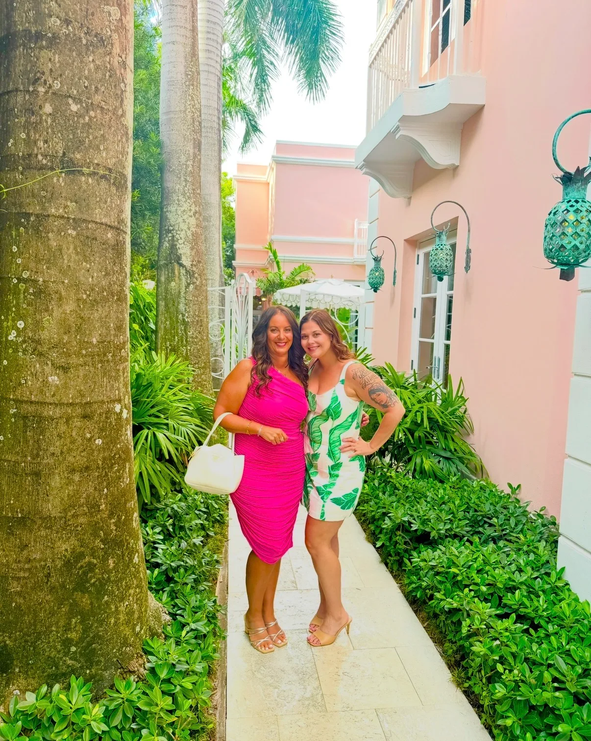 Two women pose on a garden path lined with lush tropical greenery beside a pastel pink resort building.