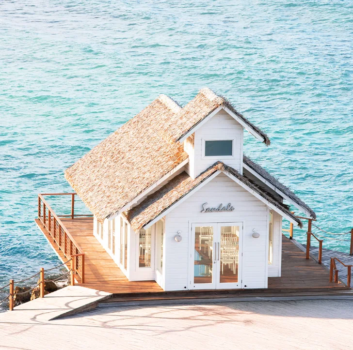 A white overwater wedding chapel with a thatched roof sits on a wooden deck above clear turquoise ocean water.