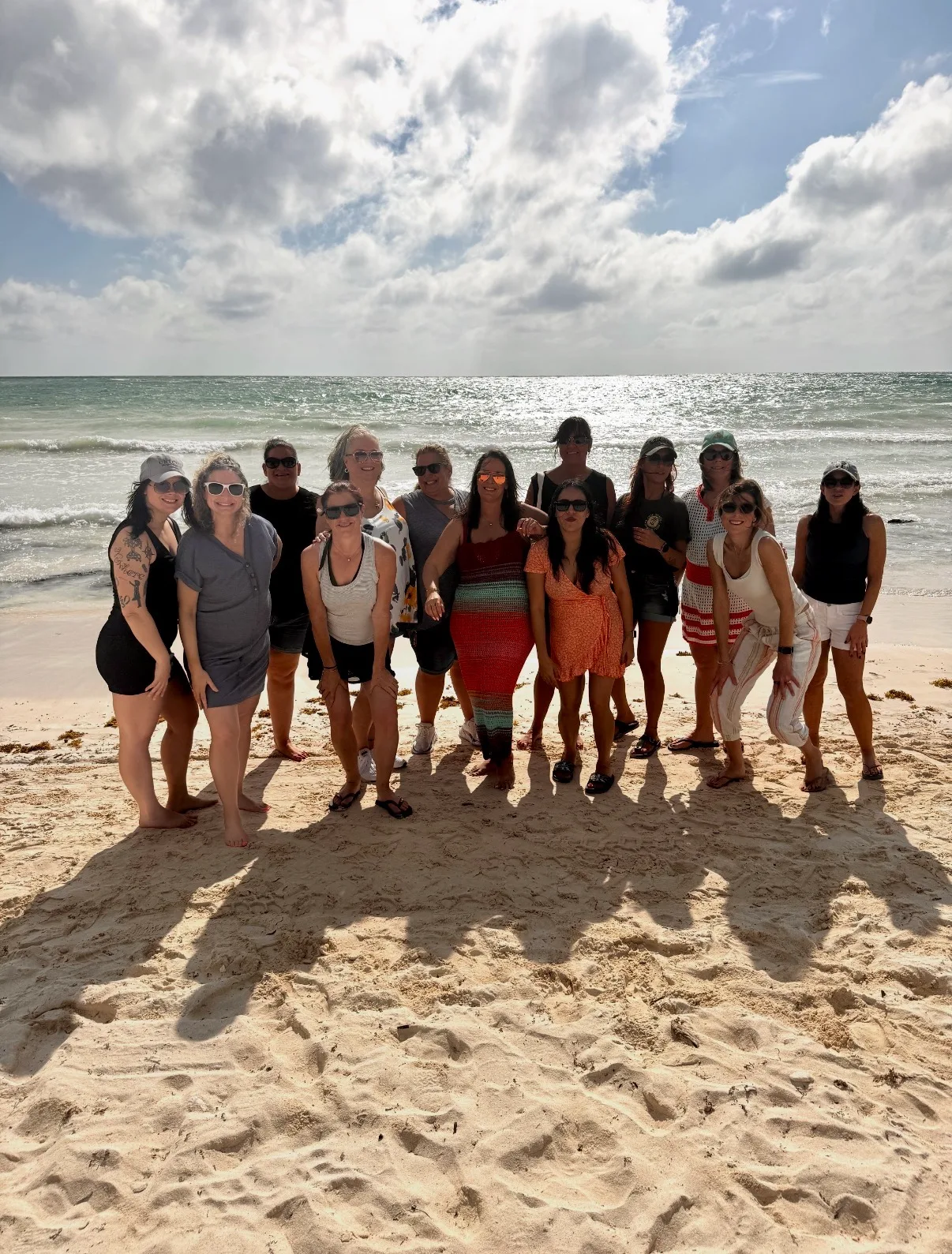 A group of women in resort wear stand together on a sandy beach before gentle ocean waves under a bright sky.