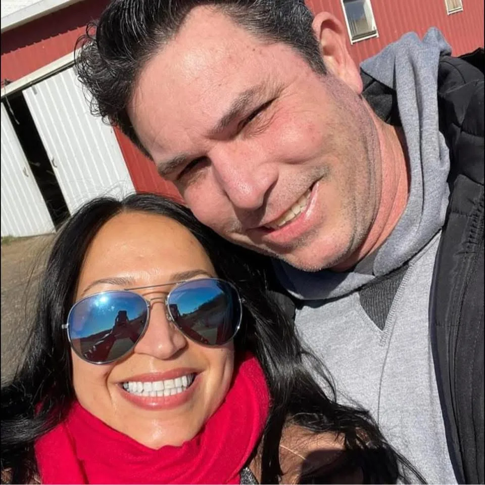 A smiling couple takes an outdoor selfie together on a sunny day in front of a red barn.