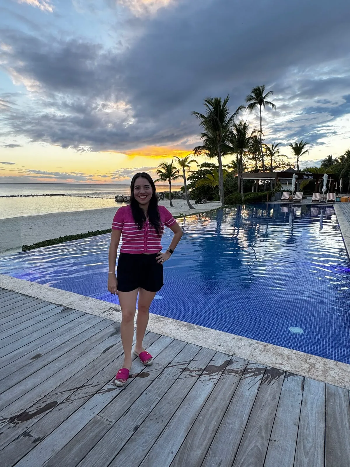 A woman stands on a wooden deck beside an infinity pool overlooking a tropical beach at sunset.