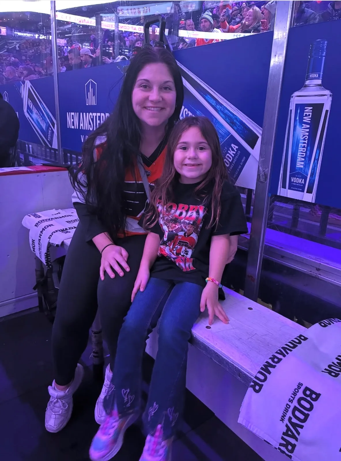 A woman and a young girl smile together while sitting on a team bench inside an ice hockey arena.