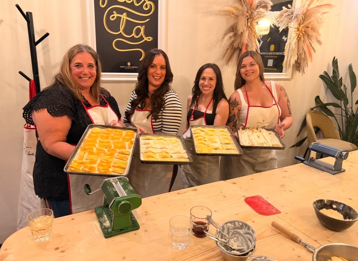 Four women wearing aprons display trays of handmade pasta behind a wooden prep counter in a Florence cooking class.