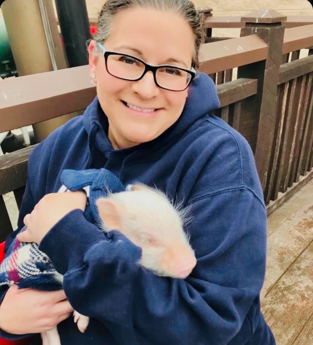 A smiling woman wearing glasses cradles a sleeping baby piglet wearing a sweater outdoors beside a wooden railing.
