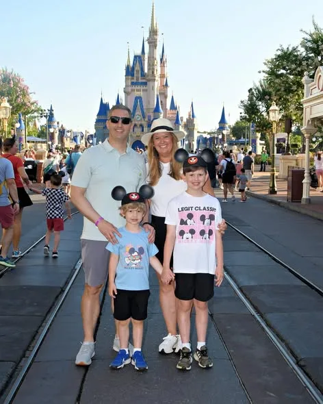 A smiling family of four wearing Mickey ears poses on Main Street in front of Cinderella Castle at Magic Kingdom.