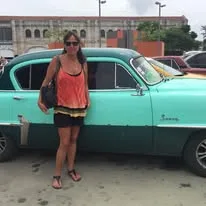 A woman in an orange top and sunglasses stands beside a vintage teal sedan parked on an urban street.