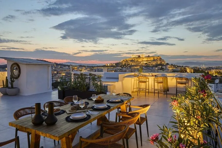 A set dining table sits on a rooftop terrace overlooking the illuminated Acropolis in Athens at sunset.