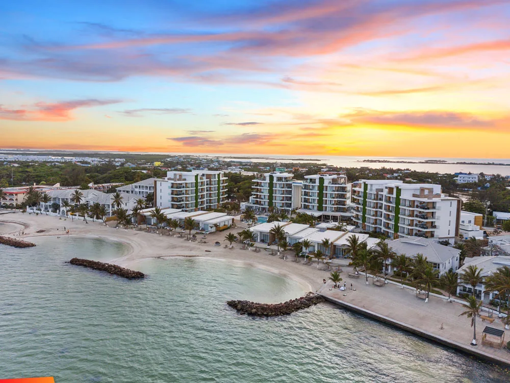 Modern beachfront resort buildings overlook sandy shores and turquoise waters beneath a vibrant sunset sky in Belize.