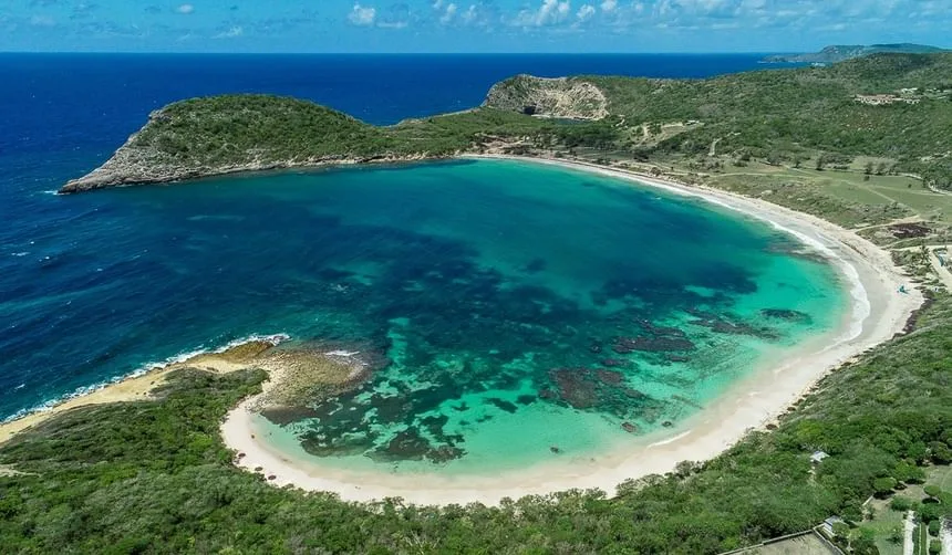 A crescent-shaped sandy beach borders turquoise Caribbean waters, framed by lush green headlands under a blue sky.