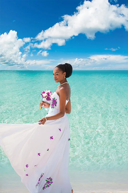 A bride in an embroidered white dress holds a bouquet against clear turquoise ocean waters under a blue sky.