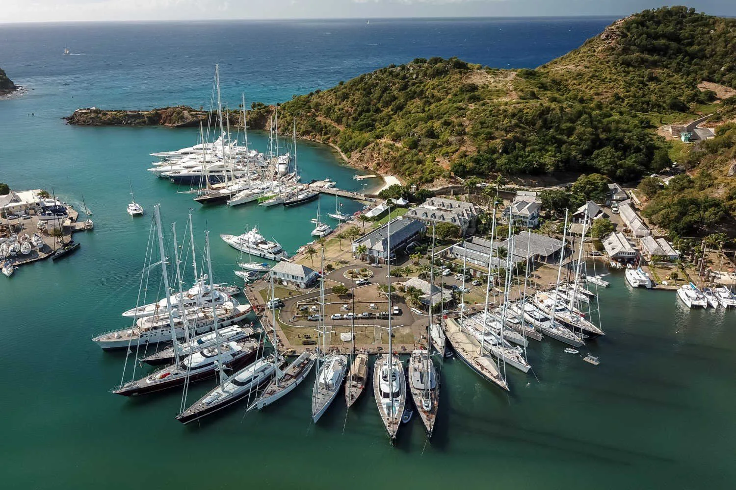 Luxury yachts and sailboats moor around a historic harbor dockyard bordered by lush green hills and turquoise Caribbean waters.