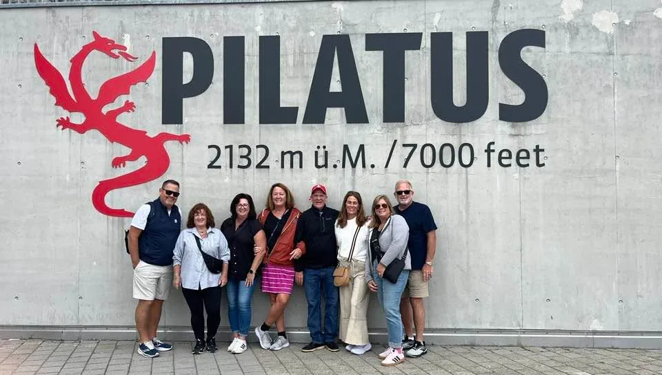 A group of travelers poses together in front of the Mount Pilatus sign and dragon logo in Switzerland.