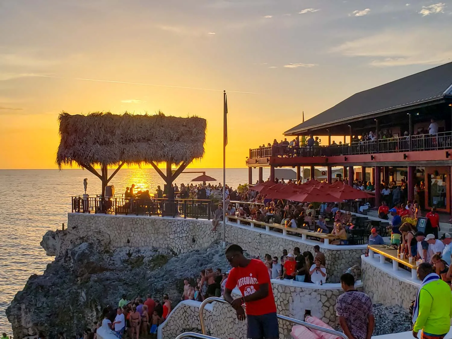 Crowds gather on the cliffside terraces of an oceanfront bar to watch the golden sunset in Negril, Jamaica.