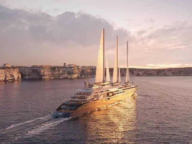 A modern three-masted sailing yacht glides along calm sea waters beside rugged coastal cliffs at sunset.