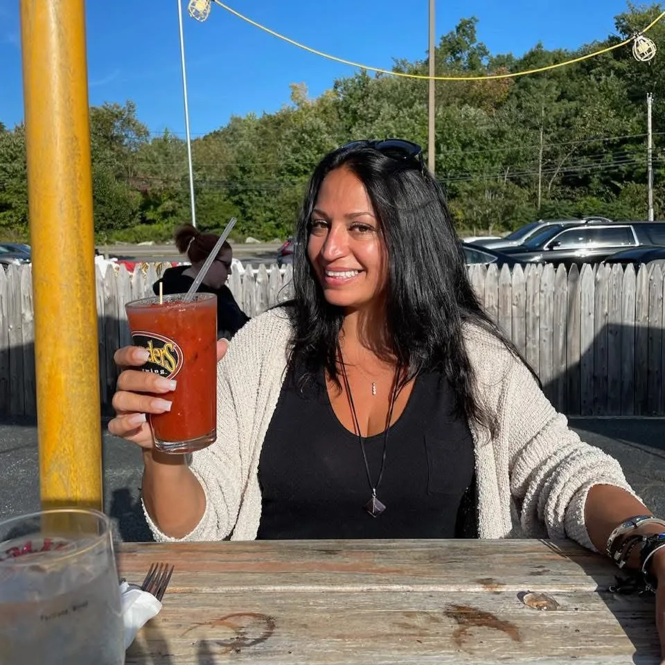 A smiling woman holds a glass with a red cocktail while sitting at a rustic outdoor patio table on a sunny day.