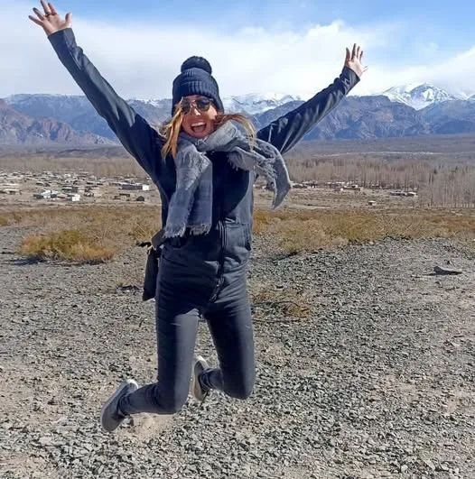 A woman wearing winter gear jumps mid-air on a rocky hillside overlooking an arid valley and distant snow-capped mountains.