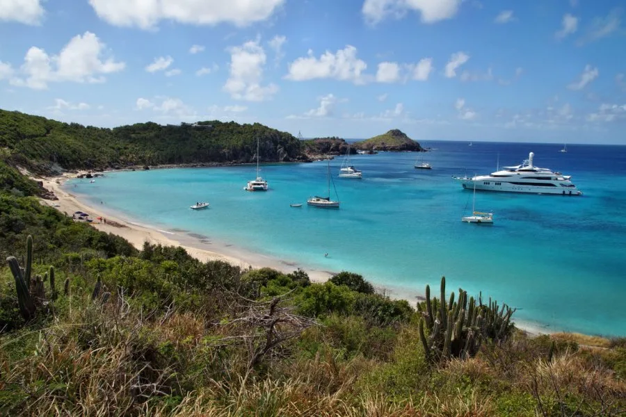 Yachts and sailboats anchor in the turquoise waters of Colombier Beach, bordered by green hills in St. Barts.