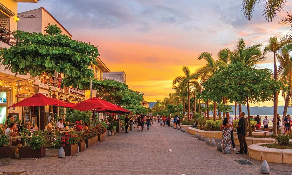 Pedestrians stroll along the palm-lined Malecón boardwalk past lively outdoor restaurants under a vibrant sunset in Puerto Vallarta, Mexico.