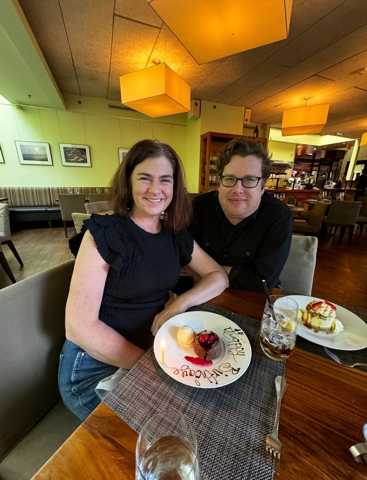 A smiling couple sits at a restaurant table with a candlelit birthday dessert plate and drinks.