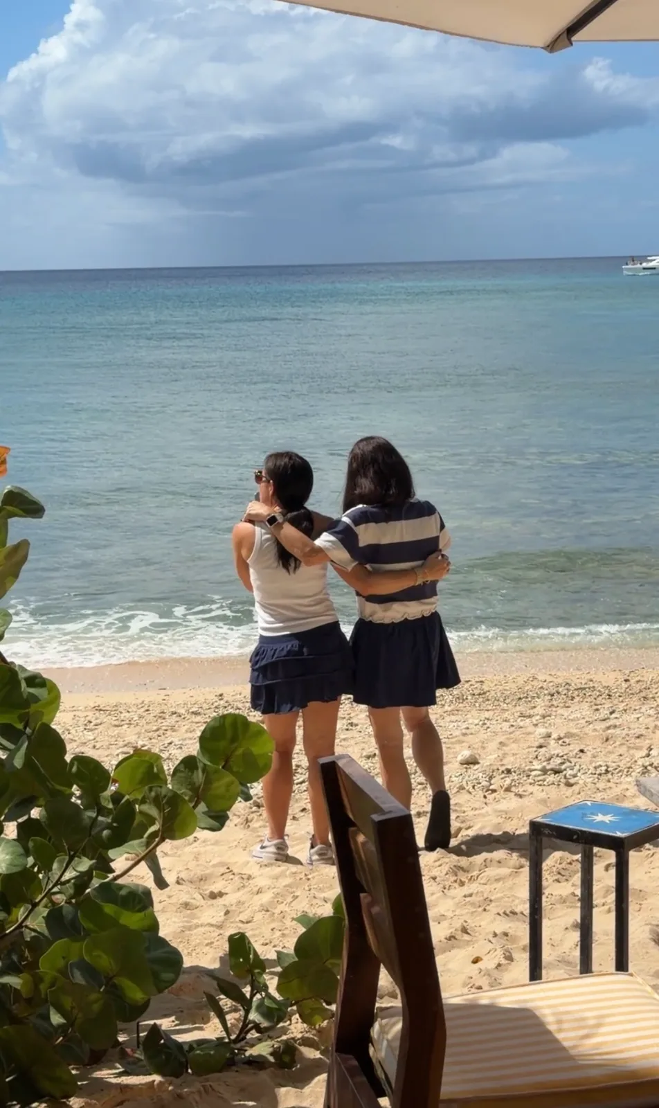 Two women embrace while looking out at the calm turquoise ocean from a sandy beach under a partly cloudy sky.