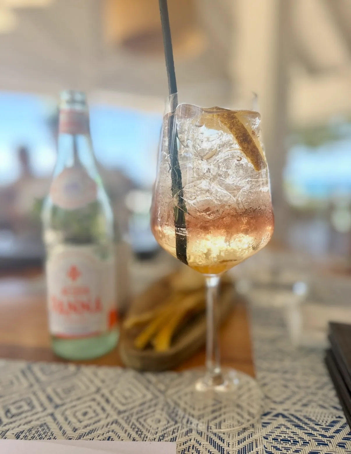 An iced cocktail with a citrus garnish sits on a table beside a bottle of mineral water at an open-air restaurant.