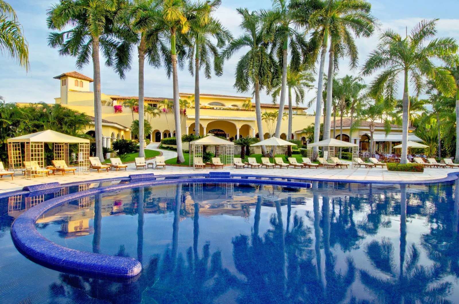 A curved blue swimming pool reflects tall palm trees outside a Puerto Vallarta resort building lined with loungers and cabanas.