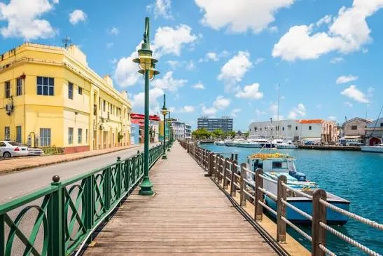 A wooden boardwalk lined with green lampposts borders a waterfront canal with moored boats and yellow buildings in Barbados.