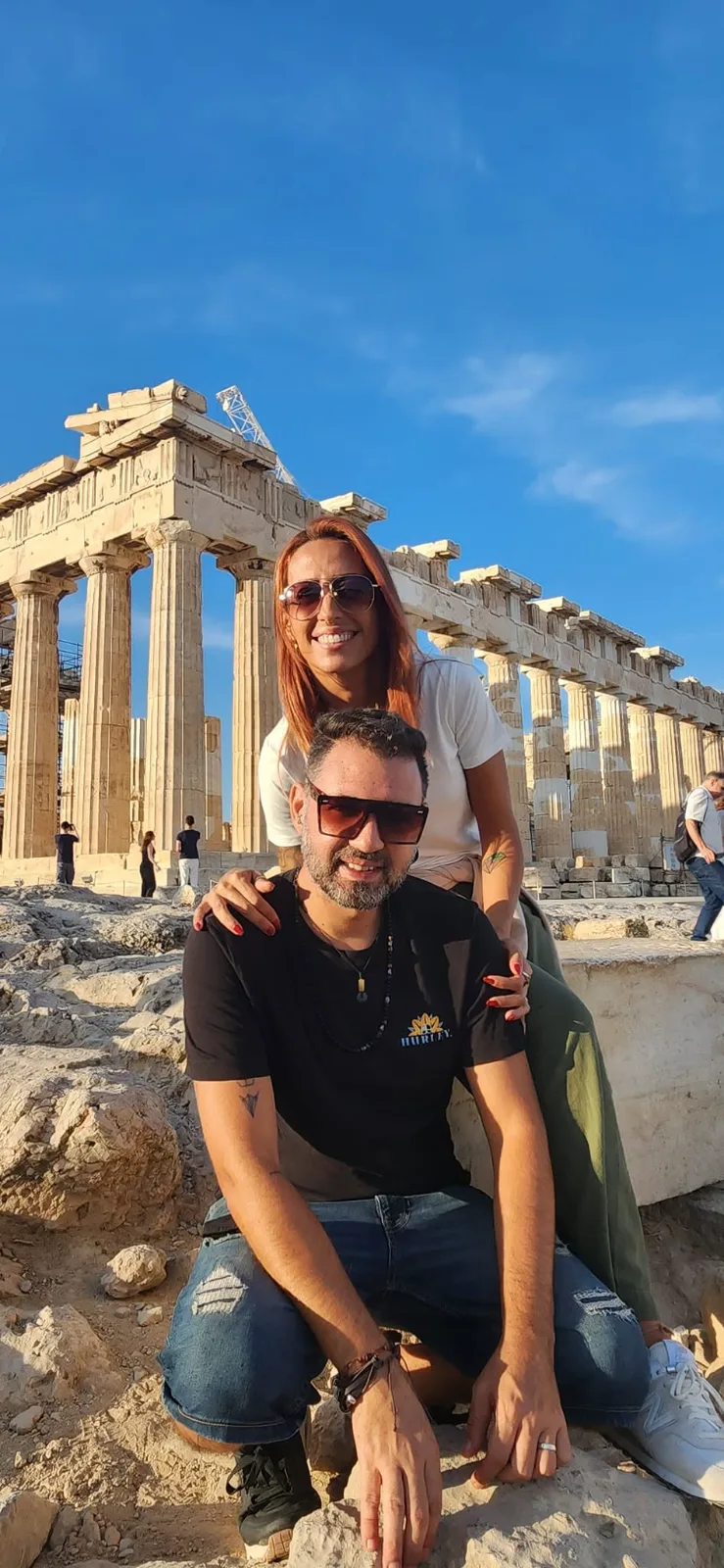 A smiling couple wearing sunglasses poses together before the ancient columns of the Parthenon in Athens, Greece.