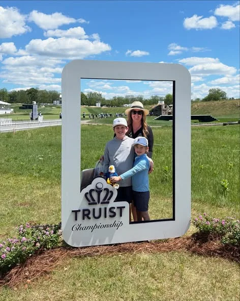 A woman and two boys pose behind a Truist Championship photo frame on a sunny golf course under blue skies.