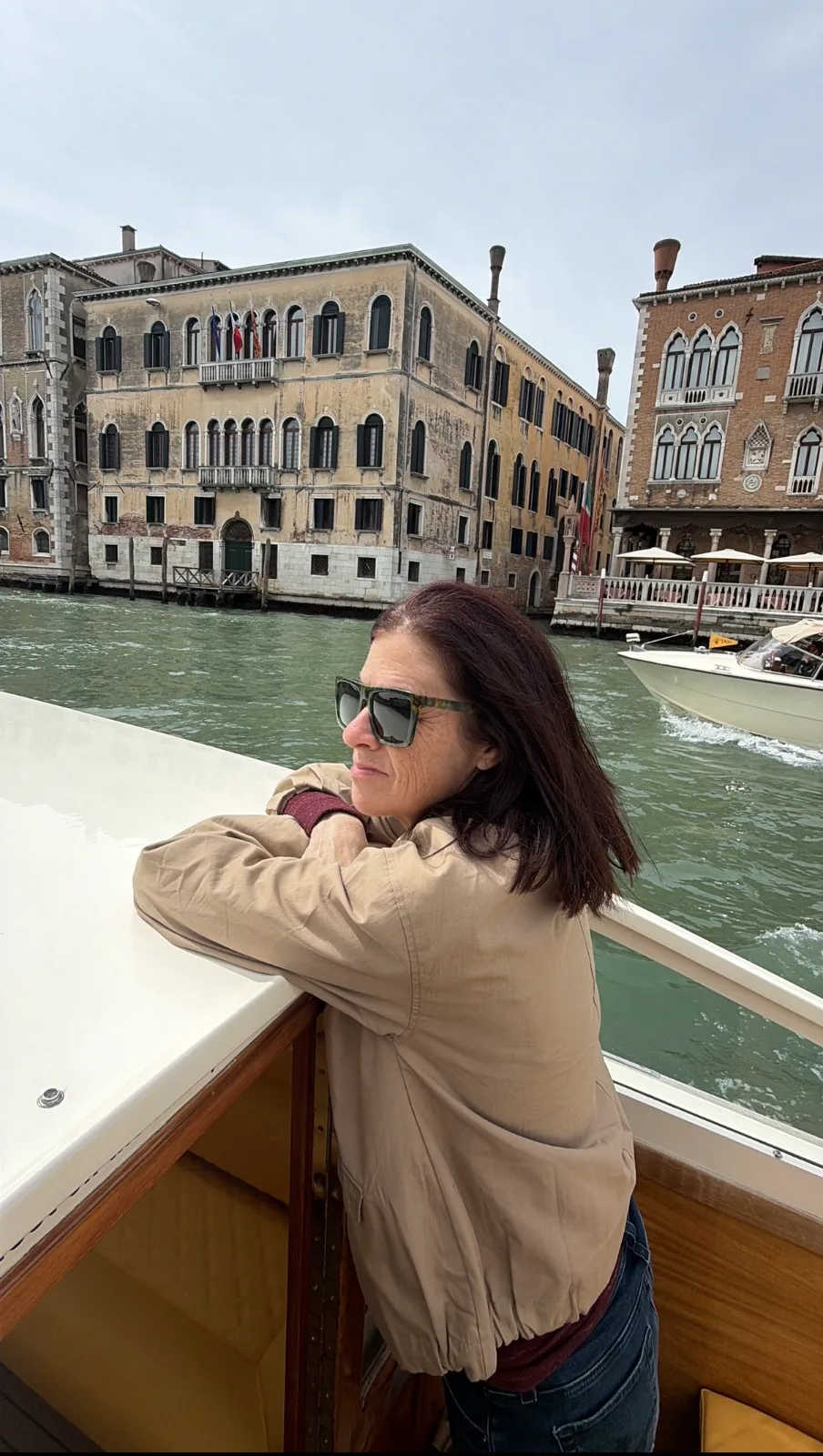 A woman in sunglasses leans against the rail of a boat cruising past historic palaces along the Grand Canal in Venice.