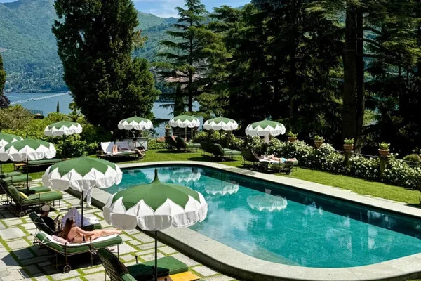 Green and white parasols shade sun loungers around an outdoor swimming pool overlooking a scenic lake and mountains in Italy.