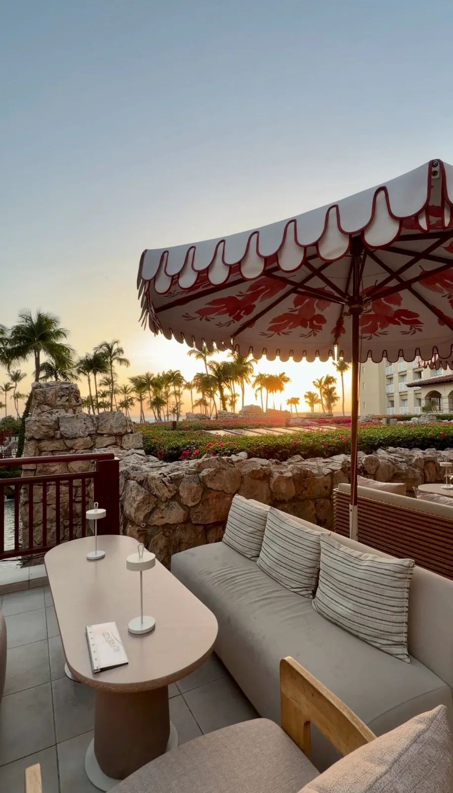 A lounge terrace with cushioned seating and a patterned umbrella overlooks palm trees at sunset in Aruba.